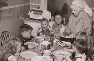 Black and white photo of VHA employee bringing food to 6 children around a dinner table