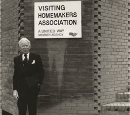 Black and white photo of man standing in front of a sign that says "visiting homemakers association"