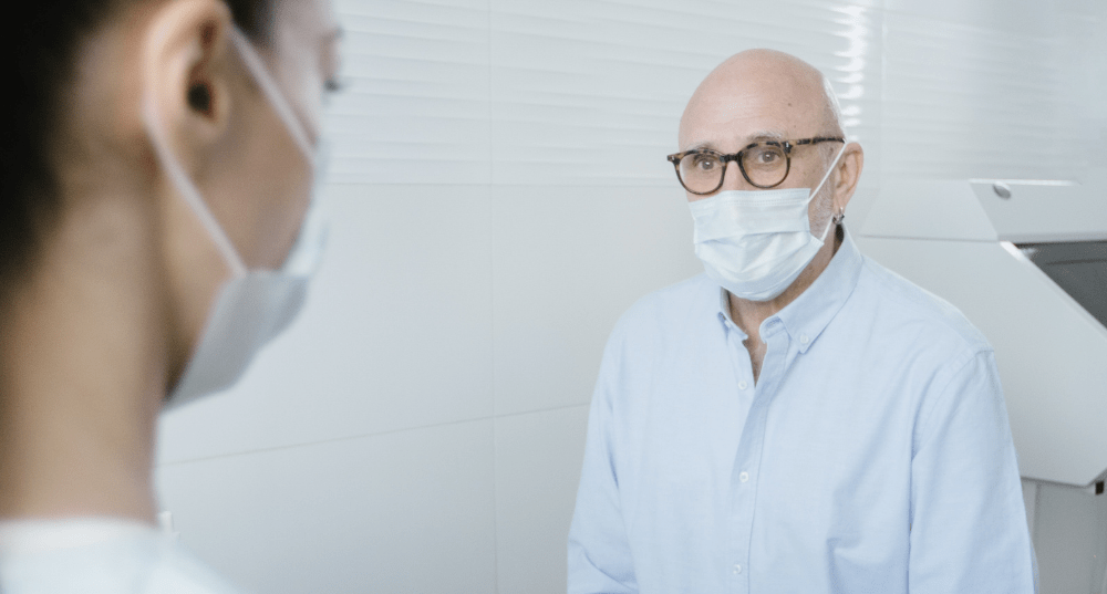 Man sits in doctor's office with his doctor while wearing a face mask