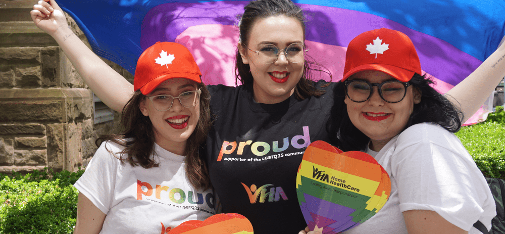 Three VHA staff members pose in front of a Pride flag during Toronto's Pride Parade