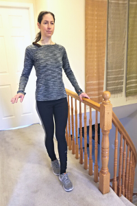 Physiotherapist walks beside a stair railing, holding the railing for balance, while walking toe-to-heel, to demonstrate the Tandem Walking balance exercise