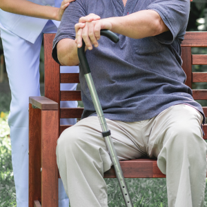 Senior east asian man sits on bench holding cane while speaking with nurse