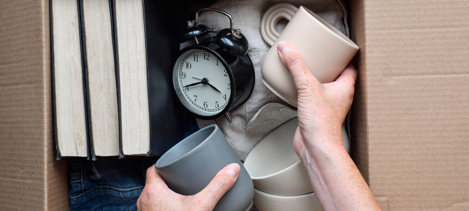Two hands placing coffee mugs into a cardboard box for storage. The box also contains other items such as an alarm clock and books.