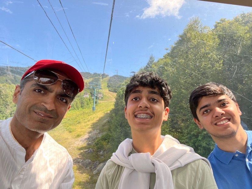 Vivek Vasudeva and his two children smiling at the camera while on a cable car. Cables and the hill in the background.