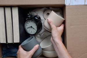 Two hands placing coffee mugs into a cardboard box for storage. The box also contains other items such as an alarm clock and books