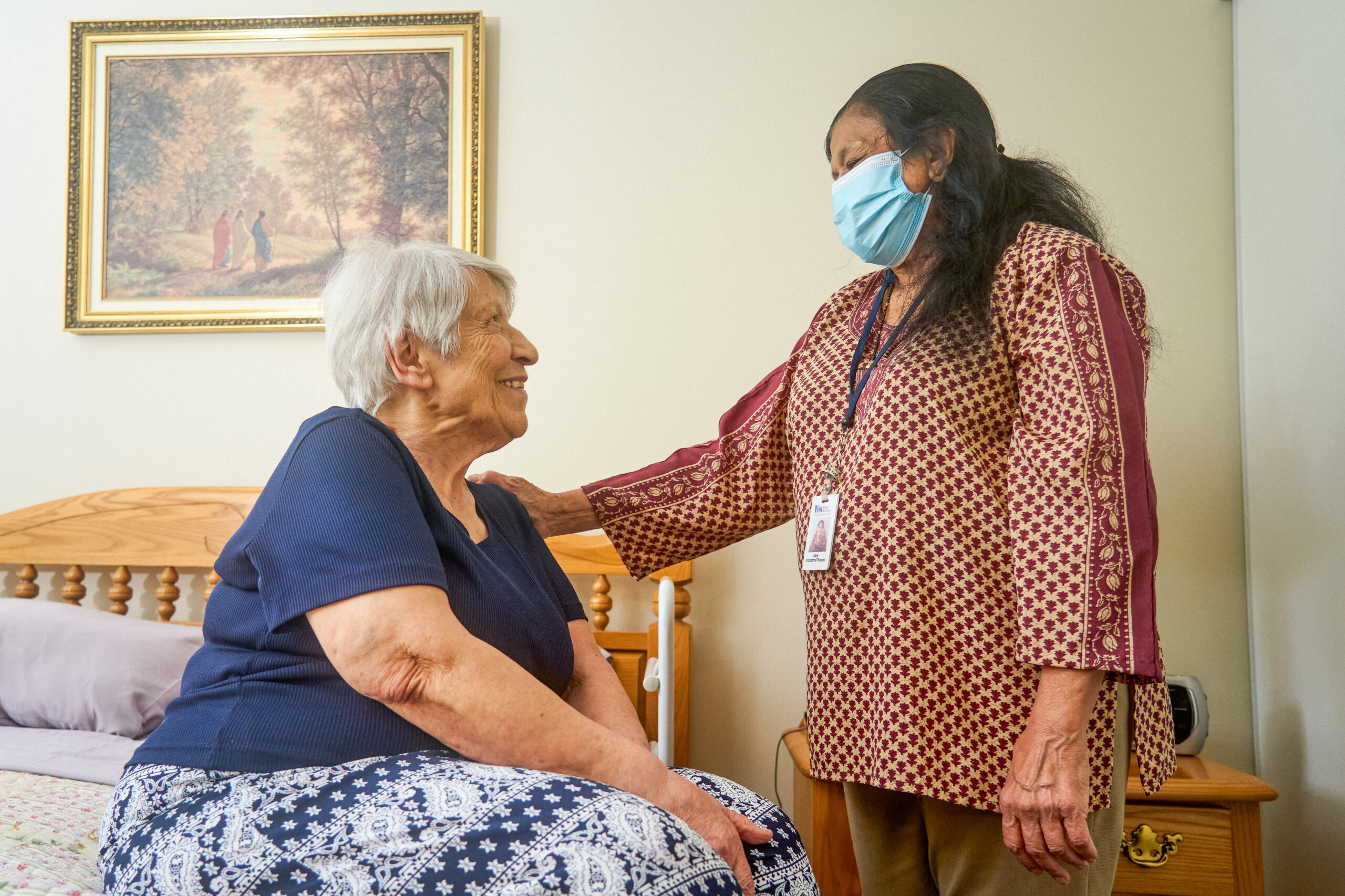 A home care provider speaking with her client. The client is sitting on the edge of her bed.