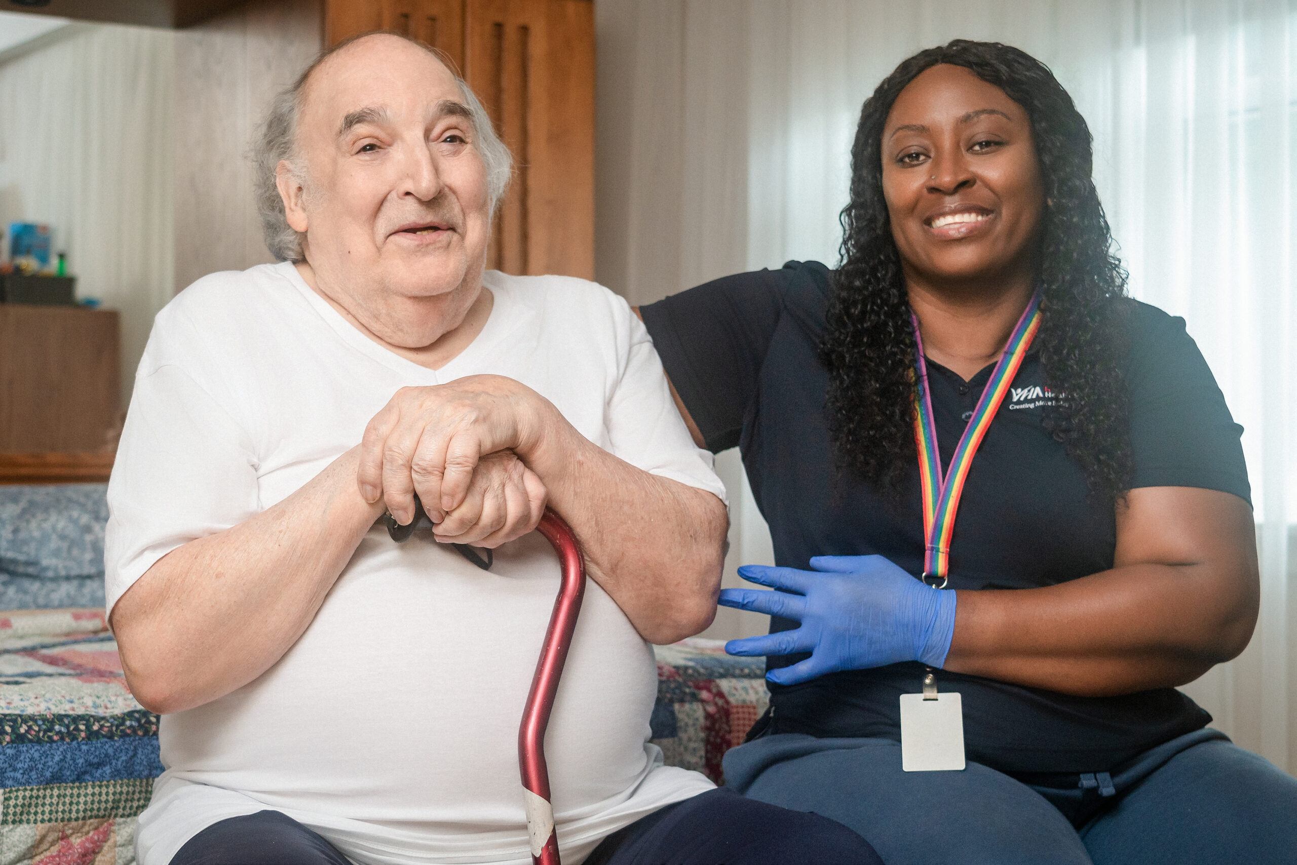 PSW, Farbiola sitting and looking at the camera with senior client, Raphael, who is holding a cane and smiling at the camera.