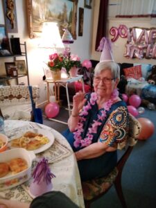 Roxanne's mother wearing a party hat, celebrates her birthday at table with food, with balloons behind her. 