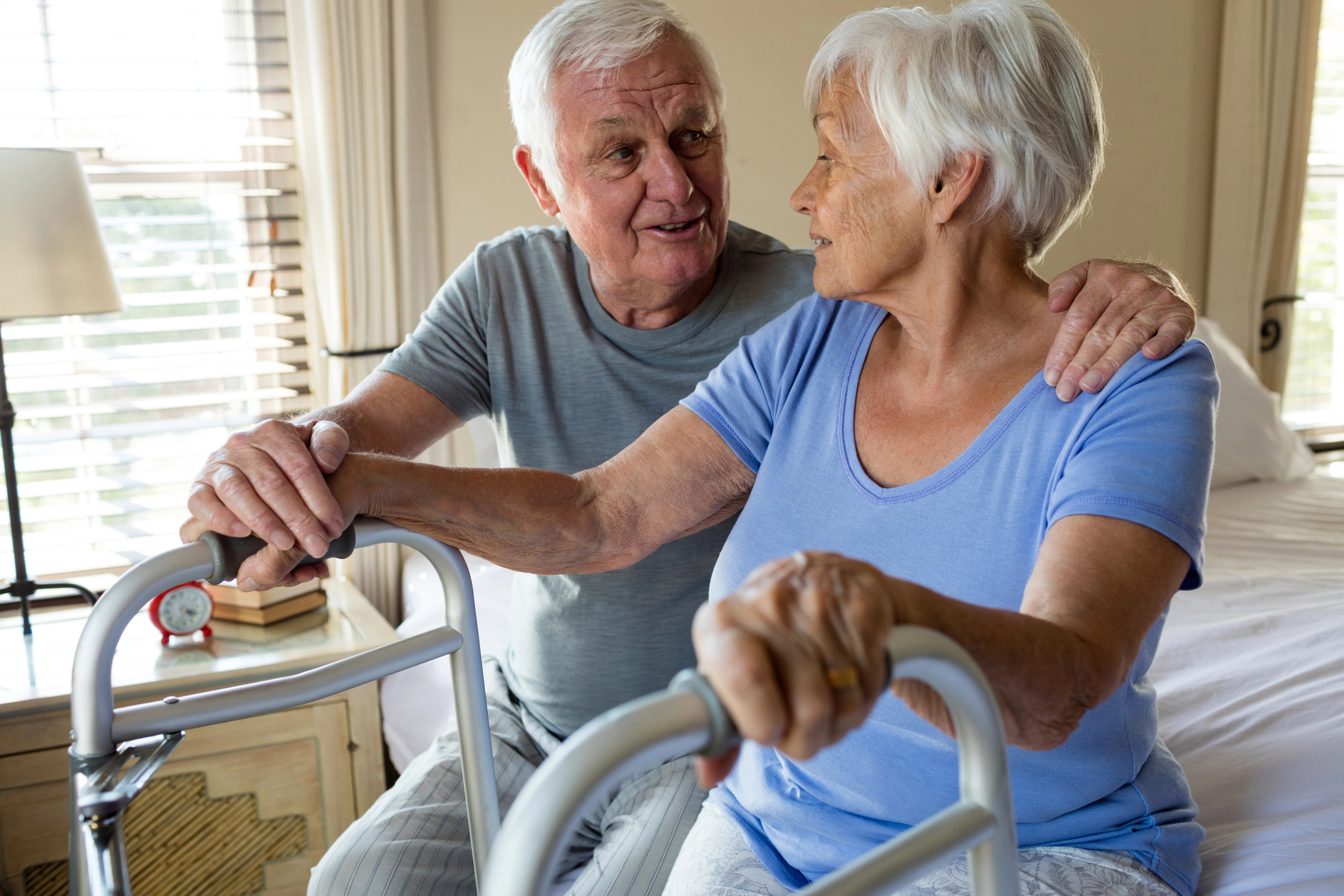 Older woman getting out of bed with a walker, with an older man supporting her with a hand on her shoulder.
